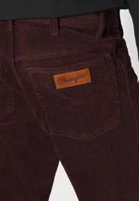 Bordo kordurojske hlače z teksturiranim vzorcem. Zadnji žep ima pravokotni usnjeni znak z izvezenim logotipom "Wrangler".