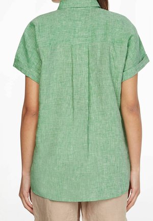 Donna che indossa una camicia verde chiaro a maniche corte con texture e schiena plissettata, abbinata a pantaloni beige, vista da dietro.