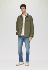 Olijfgroene lichtgewicht jas met klepzakken, gecombineerd met een geruite shirt en blauwe jeans. Witte sneakers met tan accenten maken de look compleet.
