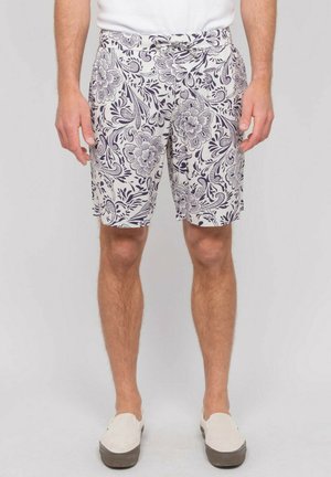 Homme portant un short blanc à motif floral noir, un t-shirt blanc uni et des chaussures blanches sans lacets, montré de la taille aux genoux, debout.