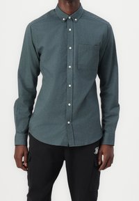 Uomo che indossa una camicia blu verde con taschino sul petto e pantaloni cargo neri con logo sulla coscia sinistra, in piedi di fronte a uno sfondo bianco.