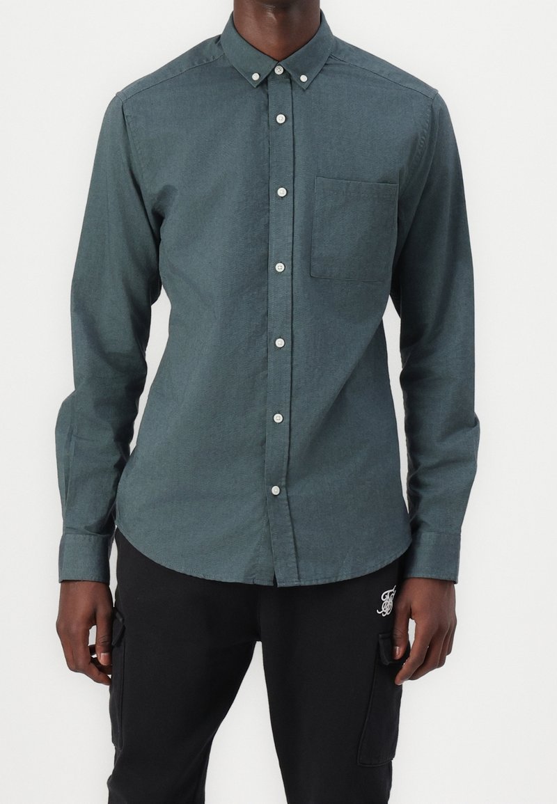 Uomo che indossa una camicia blu verde con taschino sul petto e pantaloni cargo neri con logo sulla coscia sinistra, in piedi di fronte a uno sfondo bianco.