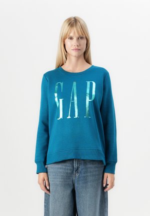 Sudadera verde azulado con cuello redondo, que presenta un gran texto metálico en plata "GAP". Tejido suave y puños acanalados. Combinada con denim claro.