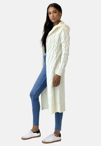 Cardigan lungo in maglia a trecce di colore verde chiaro, con cappuccio, polsini a coste e apertura frontale. Abbinato a jeans skinny e sneakers bianche.