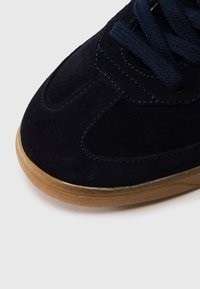 Μπλε ναυτικό σουέτ αθλητικό παπούτσι με υφασμάτινο φινίρισμα, με σόλα από καουτσούκ και κορδόνια τόνου. Διακριτικές ραφές στο πάνω μέρος. Στρογγυλεμένο σχήμα μύτης.