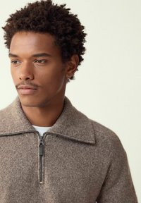 Pruun kootud pusa, millel on tõmblukuga kaelus, lai krae ja pehme tekstuur. Materjal näib soe ja mõnus, sobides ideaalselt jahedamateks ilmadeks.