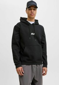 Sort hoodie i blødt stof med kengurulomme, justerbare snore og hvidt logo på brystet. Bæres med grå bukser.