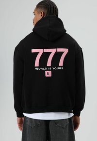 Zwarte hoodie met een grote roze "777" afbeelding en de tekst "WORLD IS YOURS" op de achterkant; zachte stof met een relaxte pasvorm.