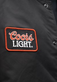 Μαύρο σακάκι από ύφασμα με ένα ορθογώνιο κεντητό έμβλημα που γράφει "Coors Light" με λευκά και πορτοκαλί γράμματα, περιγραμμένο με πορτοκαλί.