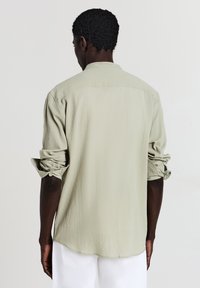 Camicia verde chiaro, button-up, a maniche lunghe, con un tessuto testurizzato, maniche arrotolate e un orlo arrotondato vista da dietro.