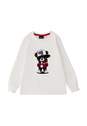 Camicia bianca a maniche lunghe con una grafica di un orsetto che indossa una giacca rossa, scarpe da ginnastica bianche e un grande cappellino da baseball bianco inclinato di lato.