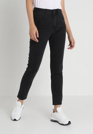 Persona vistiendo jeans negros ajustados y zapatillas blancas, de pie frente a un fondo claro y liso.