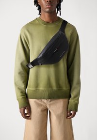 Man wearing olive green sweatshirt and beige pants with black crossbody bag featuring Tommy Hilfiger logo.