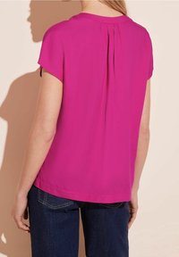 Kortärmad topp i livlig rosa med en veckdetalj på baksidan. Slätt tyg och avslappnad passform, ihop med mörka denimjeans.