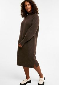 Braunes gestricktes Pulloverkleid mit hohem Kragen, langen Ärmeln, seitlichen Schlitzen und gerippten Bündchen, kombiniert mit weißen Slippern und einer schwarzen Plateausohle.