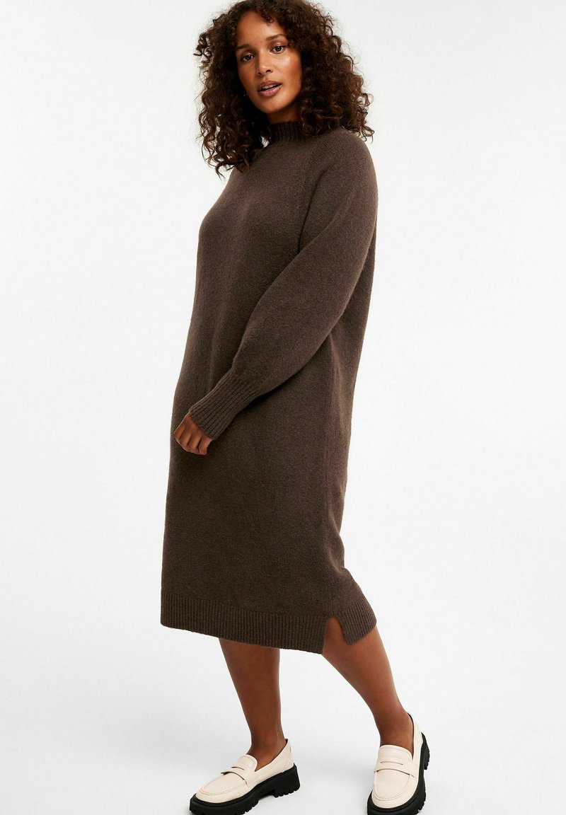 Braunes gestricktes Pulloverkleid mit hohem Kragen, langen Ärmeln, seitlichen Schlitzen und gerippten Bündchen, kombiniert mit weißen Slippern und einer schwarzen Plateausohle.