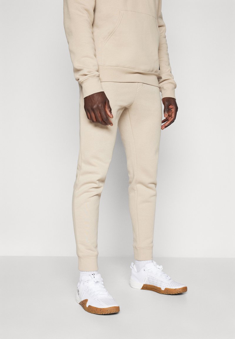Beige bomull hettegenser med kengurulomme, kombinert med beige joggebukser. Fottøyet har hvite sneakers med teksturerte brune gummisåler.