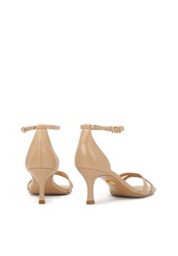 Beige nahka ankle strap -korkokengät, joissa on kaareva muotoilu, terävä varvas, kohtuullinen korko ja vaakasuorat nauhadetails. Kultaiset logovalmisteet näkyvissä.