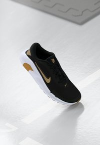 Sort og guld Nike løbesko med hvid sål, vist svævende over en reflekterende grå overflade med geometriske mønstre.