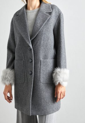 Lange grijze wollen jas met revers en drie knopen, met lichtgrijze nepbont manchetten, gedragen over een bijpassende grijze trui en broek.