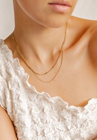 Goldfarbene Kettenhalskette mit mehreren Lagen, die über einem weißen, strukturierten Oberteil mit rüschenförmigen Kanten getragen wird, und das elegante sowie glänzende Design zur Geltung bringt.