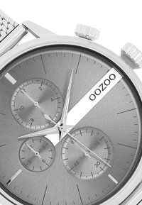Montre chronographe argentée avec un cadran gris texturé, trois sous-dials et un bracelet en maille. Présente des aiguilles des heures et des minutes nettes. Marque : OOZOO.