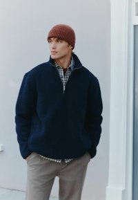 Marineblå fleece-pullover med halv lynlås og lange ærmer, lagd over en ternet skjorte. Modellen bærer en ribbet brun beanie.