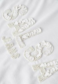 Tejido blanco con adornos texturizados en blanco formados por letras, utilizando discos y cuentas, creando un patrón decorativo.