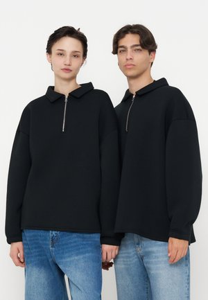 Deux personnes portant des chemises noires à manches longues avec des fermetures éclair à l'avant et des jeans bleus se tiennent côte à côte devant un fond blanc uni.