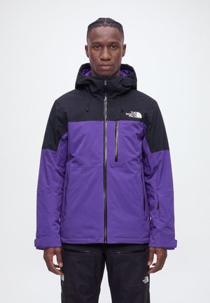 Un homme présente une veste à capuche violette et noire de The North Face avec un pantalon noir sur un fond blanc uni.