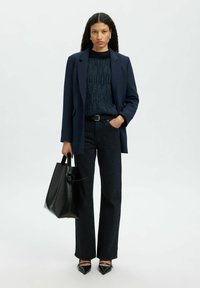 Sieviete vilkusi tumši zilu žaketi, teksturētu tumšu džemperi, melnas džinsus un smailus melnus apavus, turējusi melnu somu, stāv pie neizteiksmīgas fona.