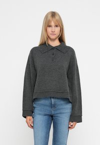 Grå kortermet genser med krage, tre knapper og brede ermer. Myk tekstur og ribbet kant, kombinert med blåjeans.