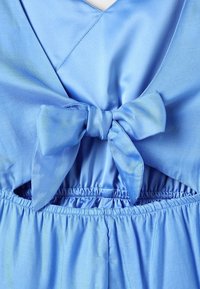 Ljust blå satin jumpsuit med v-ringning, elastisk midja och en knuten rosett på baksidan. Slät textur med lätt glans.