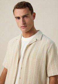 Crème shirt met korte mouwen, met witte strepen, van textuurstof en een kraag. Bevat een knoopsluiting. Draagt over een wit T-shirt.