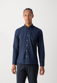Navy button-up shirt with long sleeves, made of smooth fabric, featuring a pointed collar and white buttons, paired with dark trousers.