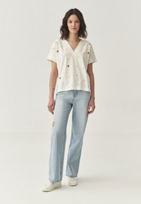 Witte V-hals top met kleurrijke, geborduurde bloemmotieven; gecombineerd met lichtblauwe straight-leg jeans met een gele bloemenpatch.