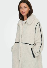 Cappotto teddy color crema, accogliente e con una texture morbida, dettagli in pipings neri, colletto ampio e tasche laterali. Presenta una chiusura con bottoni.