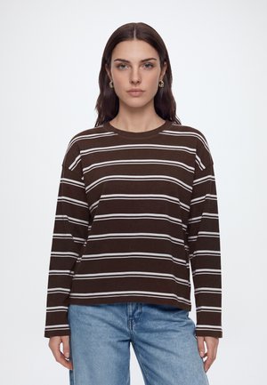 Femme aux cheveux bruns lisses portant des boucles d'oreilles en or, une chemise marron à manches longues avec des rayures horizontales blanches et un jean bleu sur un fond uni.