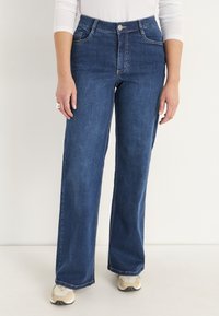 Kvinna som bär högmidjade, vida blå jeans tillsammans med beige sneakers och en vit långärmad topp, stående mot en enfärgad bakgrund.