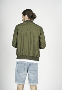Giacca bomber verde oliva con colletto e polsini a coste, in tessuto leggero e vita aderente, abbinata a pantaloni shorts in denim con bordo arrotolato.