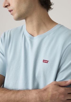 Młody mężczyzna w jasnoniebieskim T-shircie Levi's z czerwonym logo na klatce piersiowej, ze skrzyżowanymi ramionami, zwrócony w lewo na jednolitym tle.