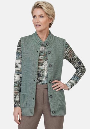 Blondharige vrouw draagt een groen gebreid mouwloos gilet met knopen over een top met lange mouwen en patroon en bruine broek, hand op de heup.
