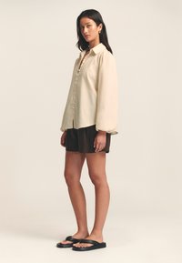 Light beige long-sleeve shirt with a button-up front, rounded hem, and slightly gathered sleeves, paired with dark shorts and black sandals.