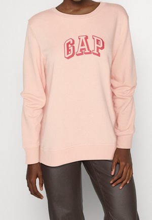 Personne portant un sweat-shirt à manches longues rose clair avec le logo rouge "GAP" sur la poitrine et un pantalon marron foncé.