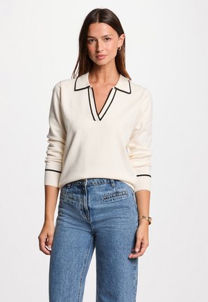 Eine Frau trägt einen cremfarbenen Pullover mit schwarzem Saum und blaue hochgeschnittene Jeans und steht vor einem schlichten weißen Hintergrund.