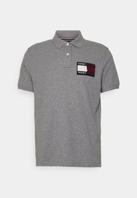 Szara koszulka polo z krótkim rękawem, z dwoma białymi guzikami oraz naszywką z logo Tommy Hilfiger i Shawna Mendesa na lewej piersi.