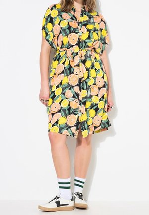 Femme portant une robe boutonnée à manches courtes avec un imprimé de fruits d’agrumes, des chaussettes blanches à rayures vertes, et des baskets blanches avec des détails noirs.