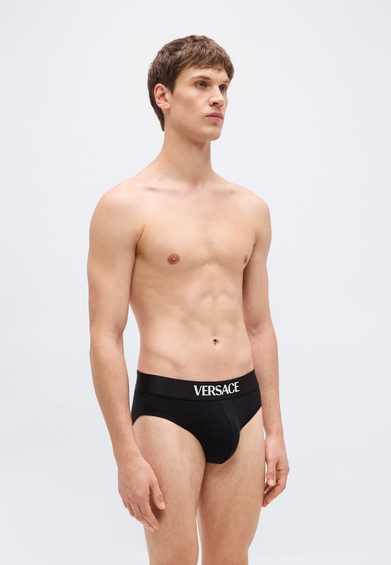 Young man standing, wearing black Versace briefs, looking slightly to the side against a plain light background.