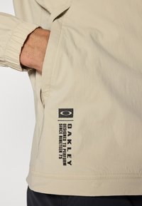 Veste d'extérieur beige avec des poignets élastiques, dotée d'une poche latérale et d'un texte de logo noir sur l'ourlet, fabriquée dans un tissu léger et texturé.