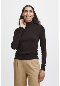 Mörkbrun polotröja, figurnära design, ribbade muddar och nederkant, tillsammans med beige byxor, visad på en leende kvinna.